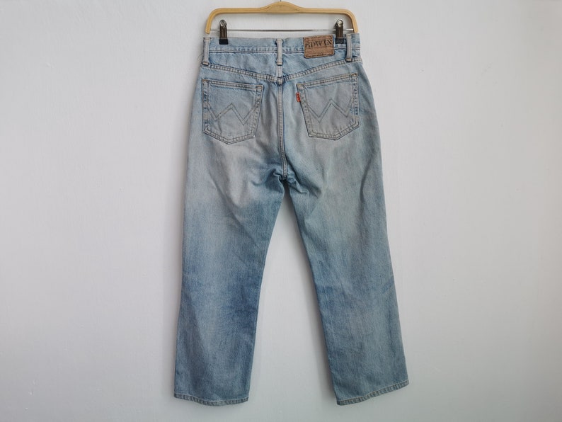 以下が含まれることがあります： 色あせたウォッシュ加工のライトブルーデニムジーンズ。Edwinブランドのラベルと赤いタグが付いています。ジーンズは、クラシックな5ポケットデザインとストレートレッグカットです。ジーンズは木製のハンガーに吊るされています。