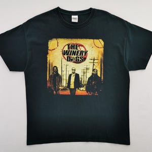 Op de afbeelding: Zwart t-shirt met een afbeelding van drie mannen op een straat en de tekst "THE WINERY DOGS" in een cirkelvormig ontwerp. De afbeelding heeft een vintage, distressed look.