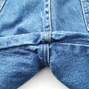 May include: Close-up of blue denim jeans, showcasing the stitching and seams. The image highlights the texture and construction of the fabric, with visible details of the thread and the overall design of the garment.