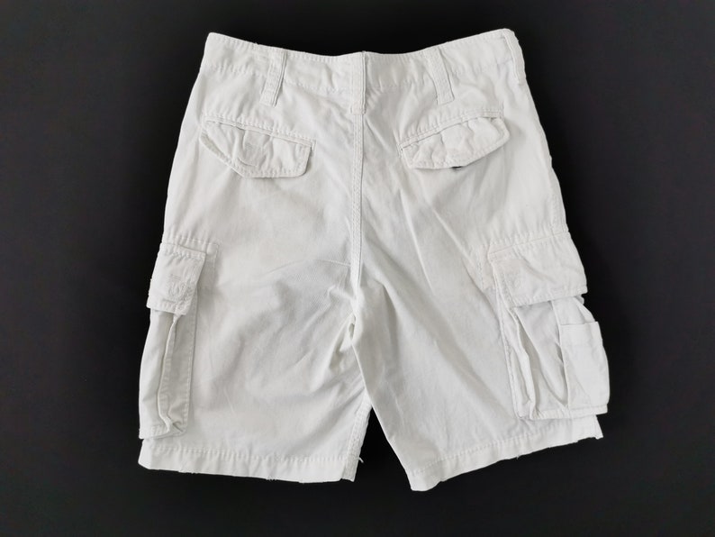 May include: White cargo shorts with multiple pockets, including two side pockets and two back pockets. The shorts have a button and zip closure and belt loops. The shorts are made of a durable fabric.