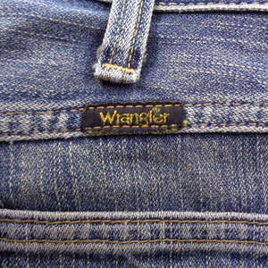 Pu&ograve; includere: Primo piano di jeans di denim blu con il marchio "Wrangler" su un'etichetta nera. I jeans presentano cuciture visibili e un design classico, che mostra la trama e i dettagli del tessuto.