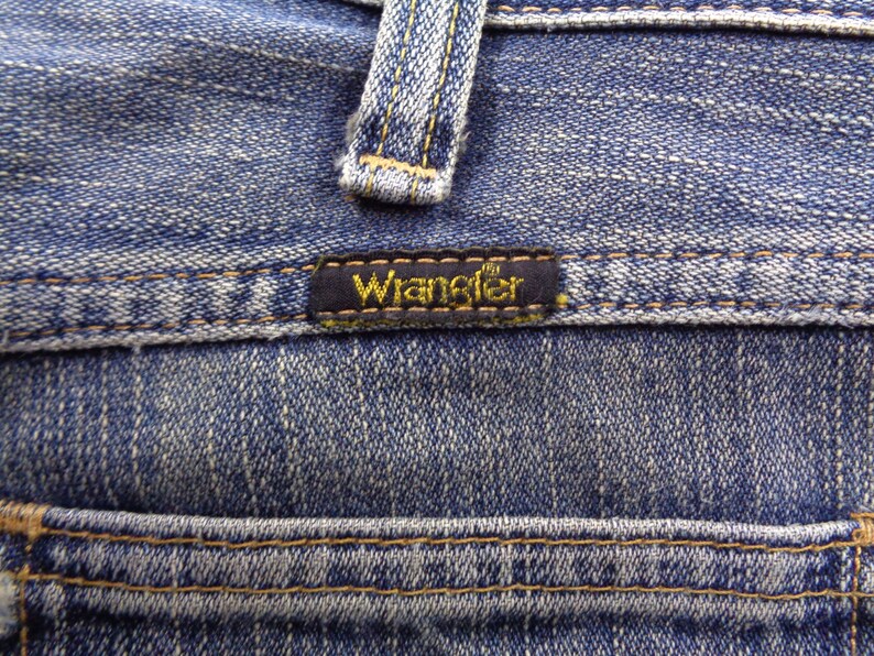 Pu&ograve; includere: Primo piano di jeans di denim blu con il marchio "Wrangler" su un'etichetta nera. I jeans presentano cuciture visibili e un design classico, che mostra la trama e i dettagli del tessuto.
