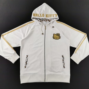 May include: White zip-up hoodie featuring a Hello Kitty graphic on the chest. The hoodie has gold stripes on the sleeves and the word "HELLO KITTY" in gold on the hood. The hood lining and pocket accents have a leopard print.