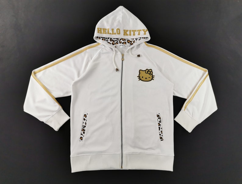 May include: White zip-up hoodie featuring a Hello Kitty graphic on the chest. The hoodie has gold stripes on the sleeves and the word "HELLO KITTY" in gold on the hood. The hood lining and pocket accents have a leopard print.
