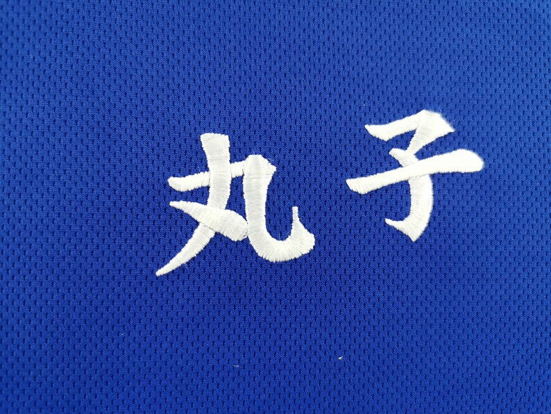 Puede incluir: Primer plano de una tela de malla azul con caracteres japoneses blancos bordados. Los caracteres est&aacute;n estilizados y parecen ser un nombre o una palabra. La tela tiene un aspecto texturizado.