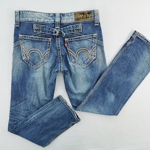 May include: Blue denim jeans with decorative stitching on the back pockets and a unique belt loop design. The jeans have a faded wash and a small red tag. The label on the back reads "Blue Trip".