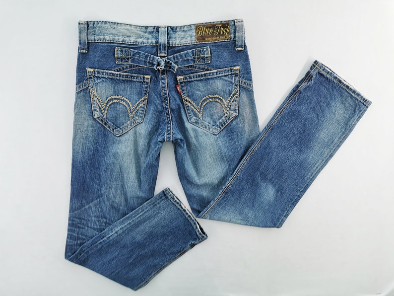 May include: Blue denim jeans with decorative stitching on the back pockets and a unique belt loop design. The jeans have a faded wash and a small red tag. The label on the back reads "Blue Trip".