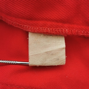 May include: Close-up of a bright red fabric with a stitched seam along the top edge. A small, rectangular, off-white tag is attached to the fabric. A silver metal object is inserted into the tag.