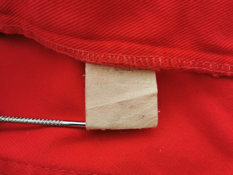 May include: Close-up of a bright red fabric with a stitched seam along the top edge. A small, rectangular, off-white tag is attached to the fabric. A silver metal object is inserted into the tag.