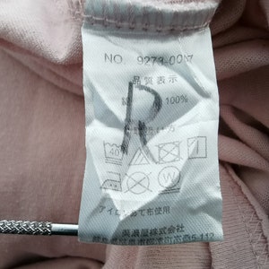 May include: A close-up of a pink fabric tag with Japanese text and care symbols. The tag includes the number "9279-007" and the text "100%" on a pink garment.