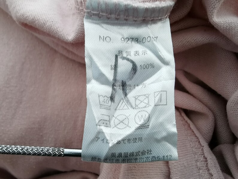 May include: A close-up of a pink fabric tag with Japanese text and care symbols. The tag includes the number "9279-007" and the text "100%" on a pink garment.