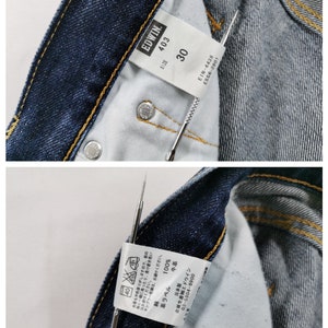 May include: Close-up of the inside of blue denim jeans. The jeans have a visible tag with the brand "EDWIN" and size "30". The tag also includes washing instructions and other details. The jeans have a classic five-pocket design.