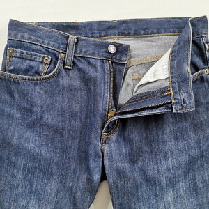 May include: Close-up of blue denim jeans with a partially unzipped fly. The jeans have a classic five-pocket design, with visible stitching and metal rivets. The fabric shows a slight fade, indicating wear.