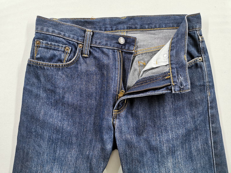 May include: Close-up of blue denim jeans with a partially unzipped fly. The jeans have a classic five-pocket design, with visible stitching and metal rivets. The fabric shows a slight fade, indicating wear.
