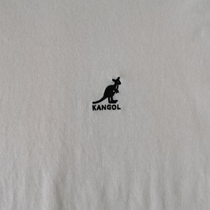 Op de afbeelding: Cr&egrave;mekleurige stof met een zwart kangoeroelogo en de merknaam "KANGOL" eronder gedrukt. Het logo is een silhouet van een kangoeroe. De stof heeft een subtiele textuur.