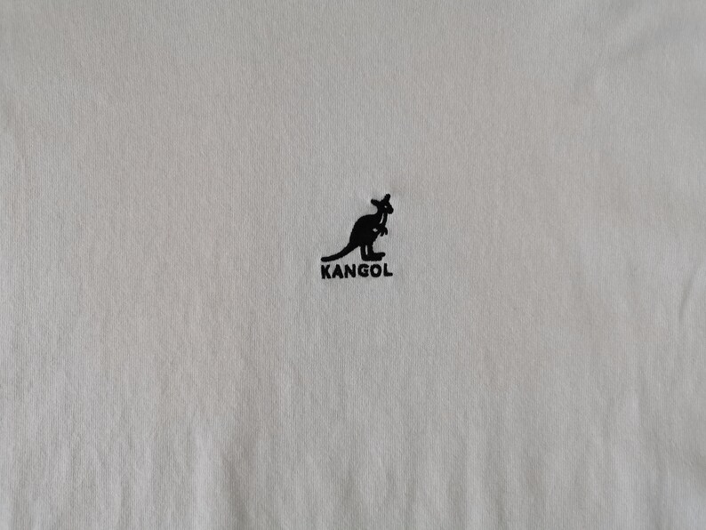 Op de afbeelding: Cr&egrave;mekleurige stof met een zwart kangoeroelogo en de merknaam "KANGOL" eronder gedrukt. Het logo is een silhouet van een kangoeroe. De stof heeft een subtiele textuur.