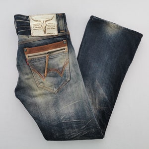 Pu&ograve; includere: Jeans blu in denim con lavaggio effetto used. Presentano un dettaglio di tasca in pelle marrone con cerniera, una toppa in pelle con un teschio di toro e cuciture rosse sulla tasca posteriore. I jeans hanno un'apertura della gamba a zampa d'elefante.
