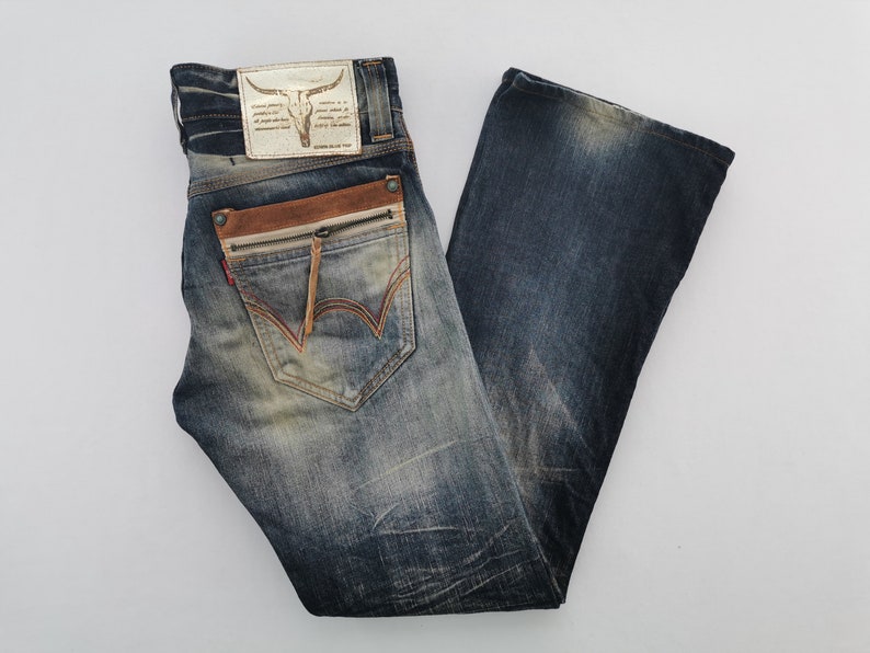 Pu&ograve; includere: Jeans blu in denim con lavaggio effetto used. Presentano un dettaglio di tasca in pelle marrone con cerniera, una toppa in pelle con un teschio di toro e cuciture rosse sulla tasca posteriore. I jeans hanno un'apertura della gamba a zampa d'elefante.