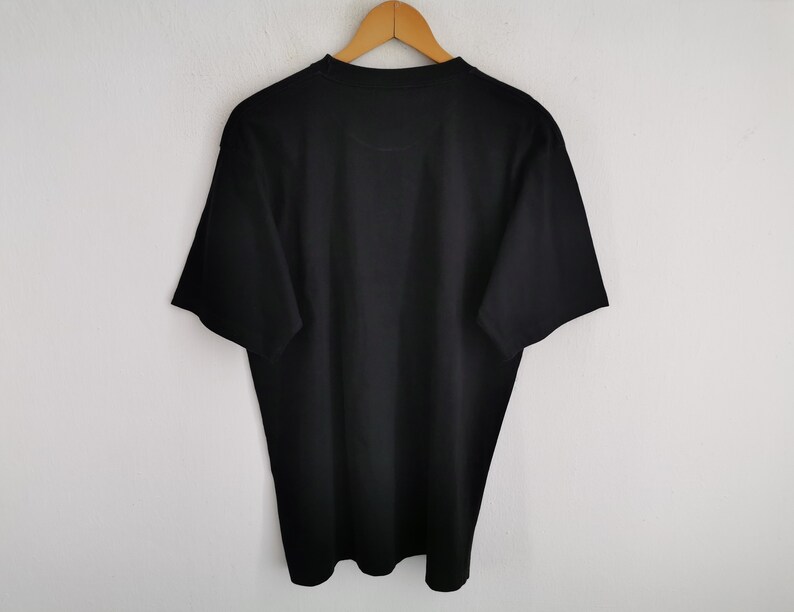 May include: A solid black t-shirt hanging on a wooden hanger against a white wall. The short-sleeved shirt is made of a soft material and has a crew neck. The shirt is a basic, everyday garment.