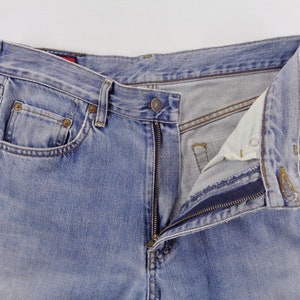 May include: Light blue denim jeans with a partially unzipped fly. The jeans have classic five-pocket styling, with rivets and a button closure. The fabric shows signs of wear, with a faded appearance.