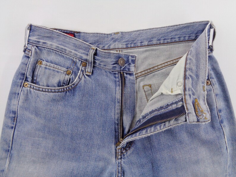 May include: Light blue denim jeans with a partially unzipped fly. The jeans have classic five-pocket styling, with rivets and a button closure. The fabric shows signs of wear, with a faded appearance.