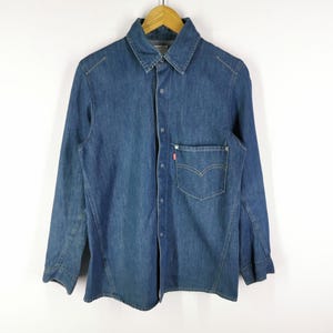 Puede incluir: Camisa vaquera de manga larga en un lavado azul medio, con cuello clásico, cierre de botones y un bolsillo en el pecho con una etiqueta con el logo rojo. La camisa está colgada en una percha de madera.