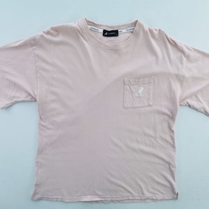 May include: A light pink t-shirt with a pocket on the chest. The pocket features a white logo of a kangaroo. The brand name is printed on the collar. The shirt is made of a soft material.