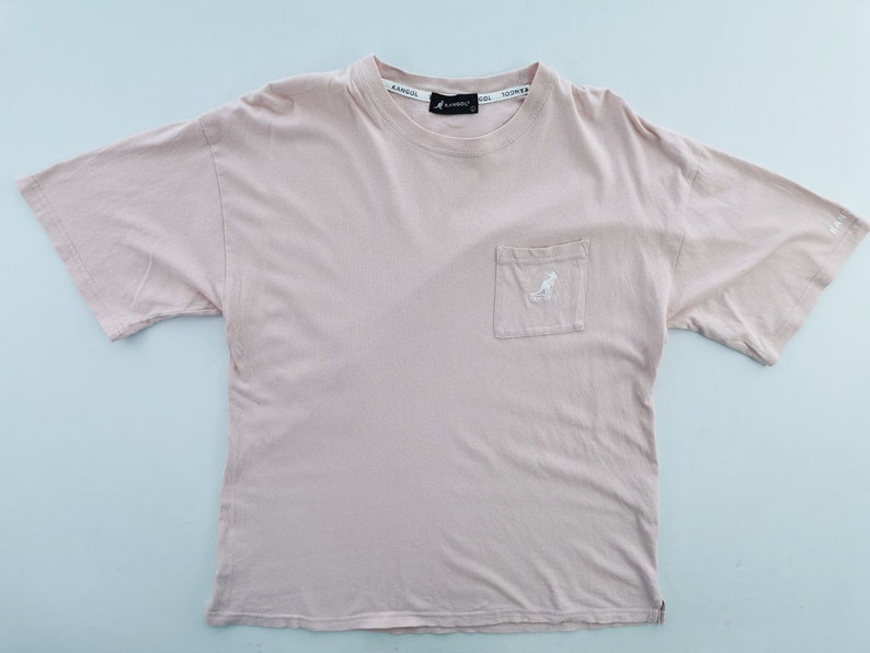 May include: A light pink t-shirt with a pocket on the chest. The pocket features a white logo of a kangaroo. The brand name is printed on the collar. The shirt is made of a soft material.