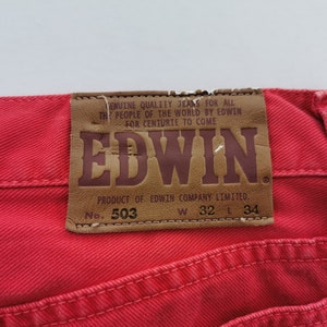 Op de afbeelding: Close-up van een rode spijkerbroek met een leren patch. De patch toont de merknaam "EDWIN" en tekst. De jeans heeft een taillemaat van 81 cm en een lengte van 86 cm.