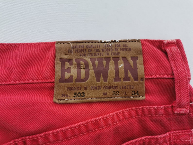 Op de afbeelding: Close-up van een rode spijkerbroek met een leren patch. De patch toont de merknaam "EDWIN" en tekst. De jeans heeft een taillemaat van 81 cm en een lengte van 86 cm.
