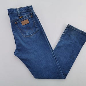 May include: A pair of medium-wash blue denim jeans featuring a high-waisted cut, a classic five-pocket design, and a brown leather patch on the back pocket with the Wrangler brand name printed in black text, along with copper-colored metal rivets.