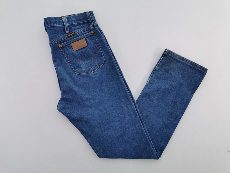 May include: A pair of medium-wash blue denim jeans featuring a high-waisted cut, a classic five-pocket design, and a brown leather patch on the back pocket with the Wrangler brand name printed in black text, along with copper-colored metal rivets.