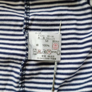 Puede incluir: Primer plano de una camisa a rayas azul marino y blanca. Se adjunta una etiqueta con texto japon&eacute;s y s&iacute;mbolos de cuidado. La etiqueta incluye informaci&oacute;n sobre la talla, medidas de pecho y altura, y la composici&oacute;n del material.