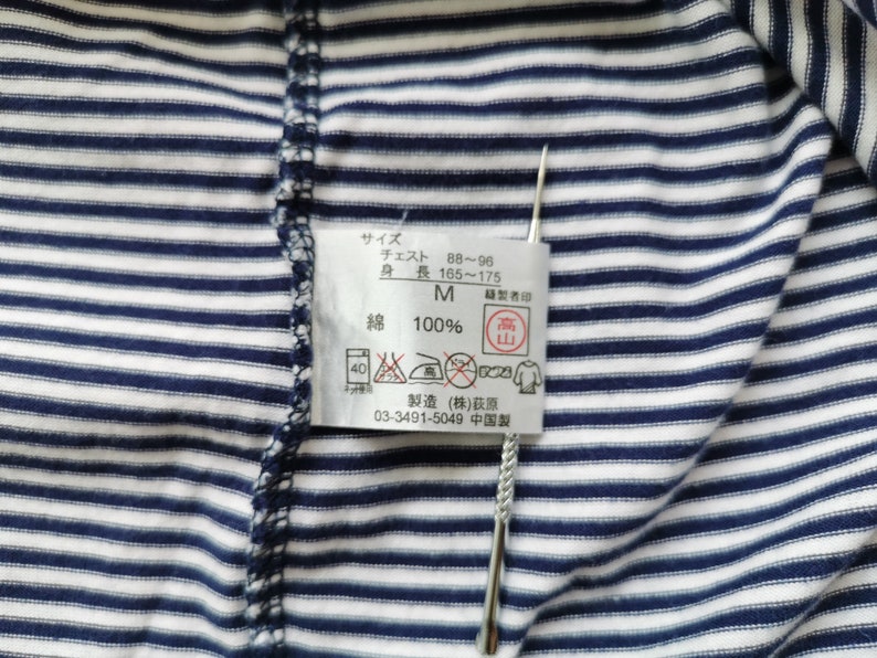 Puede incluir: Primer plano de una camisa a rayas azul marino y blanca. Se adjunta una etiqueta con texto japon&eacute;s y s&iacute;mbolos de cuidado. La etiqueta incluye informaci&oacute;n sobre la talla, medidas de pecho y altura, y la composici&oacute;n del material.