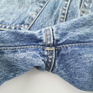 Puede incluir: Primer plano de unos vaqueros de mezclilla azul claro. La imagen destaca los detalles de la tela, las costuras y los pespuntes. Los vaqueros tienen un dise&ntilde;o cl&aacute;sico con costuras amarillas en contraste.