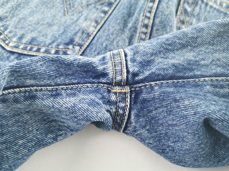 Puede incluir: Primer plano de unos vaqueros de mezclilla azul claro. La imagen destaca los detalles de la tela, las costuras y los pespuntes. Los vaqueros tienen un dise&ntilde;o cl&aacute;sico con costuras amarillas en contraste.