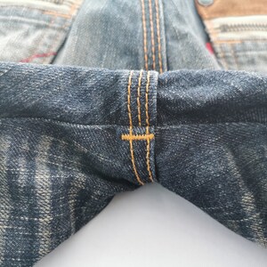 Pu&ograve; includere: Primo piano di jeans in denim con cuciture visibili in filo arancione. I jeans hanno un aspetto sbiadito e usurato con un effetto di lavaggio chiaro. Il tessuto mostra segni di usura.