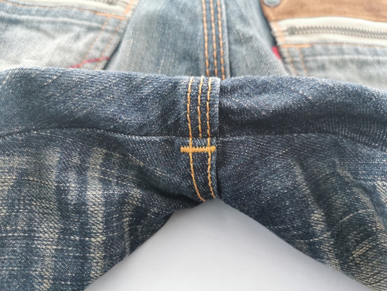 Pu&ograve; includere: Primo piano di jeans in denim con cuciture visibili in filo arancione. I jeans hanno un aspetto sbiadito e usurato con un effetto di lavaggio chiaro. Il tessuto mostra segni di usura.