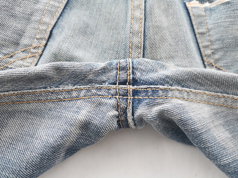 K&ouml;nnte beinhalten: Nahaufnahme einer hellblauen Jeans. Das Bild zeigt die Nahtdetails, mit sichtbaren N&auml;hten und einem kreuzgen&auml;hten Bereich. Der Denim hat ein abgenutztes Aussehen.