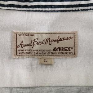 May include: A white shirt with a collar, featuring a label with the text "Armed Forces Manufacturer" and "AVIREX&reg;". The label also includes the text "AUTHENTIC GARMENT ESTABLISHED IN 1975" and the size "L".
