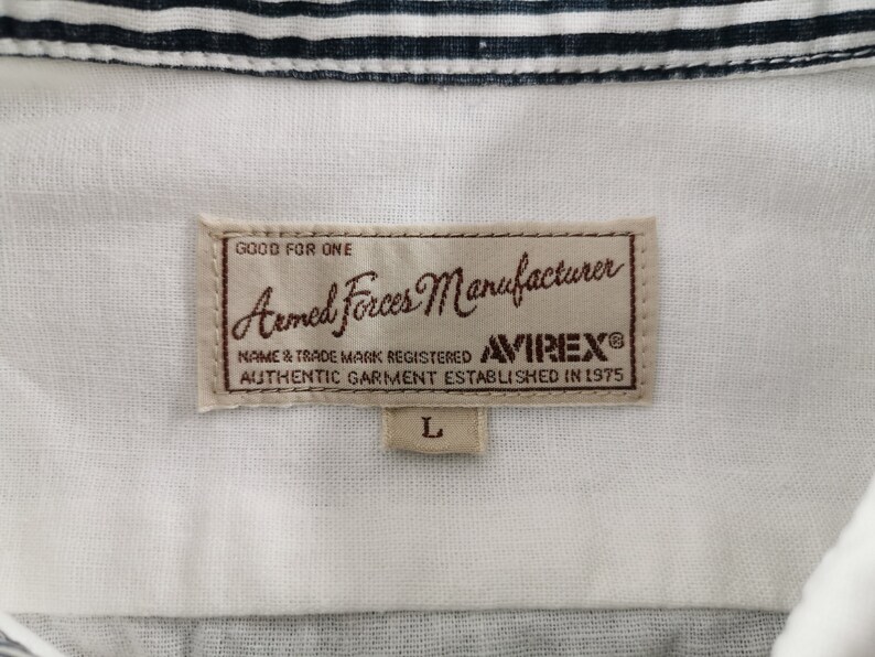 May include: A white shirt with a collar, featuring a label with the text "Armed Forces Manufacturer" and "AVIREX&reg;". The label also includes the text "AUTHENTIC GARMENT ESTABLISHED IN 1975" and the size "L".