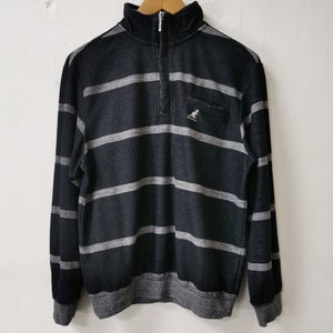 May include: Black and gray striped long-sleeve sweatshirt with a quarter-zip closure and a small pocket on the chest. The sweatshirt features the Kangol logo and a ribbed collar, cuffs, and hem.