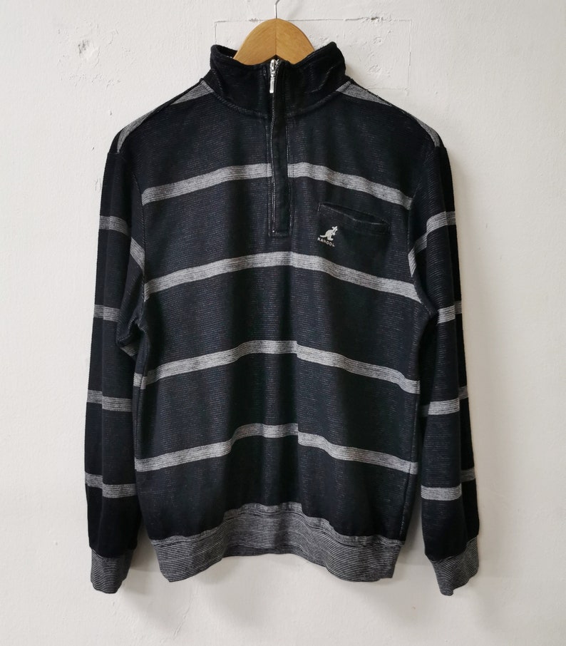 May include: Black and gray striped long-sleeve sweatshirt with a quarter-zip closure and a small pocket on the chest. The sweatshirt features the Kangol logo and a ribbed collar, cuffs, and hem.