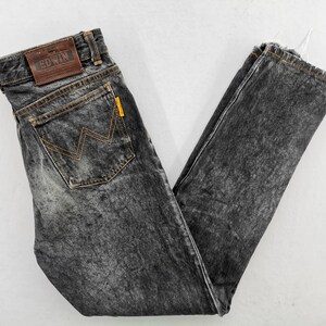 May include: Acid-wash black denim jeans featuring a brown leather patch on the waistband with the text EDWIN embossed. The back pocket displays a signature orange zigzag stitch pattern and a small yellow tab. The hem has a frayed, raw edge finish.
