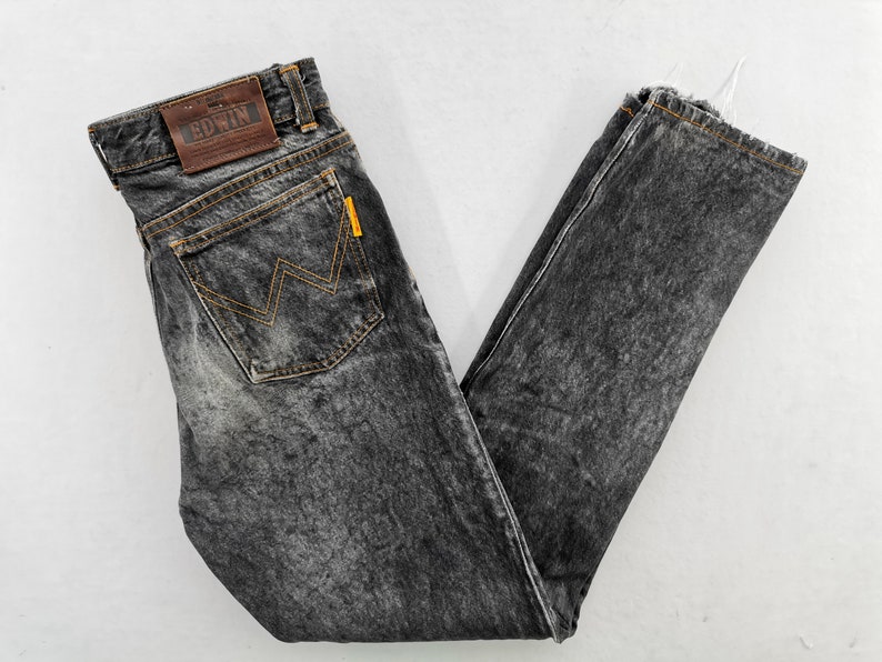 May include: Acid-wash black denim jeans featuring a brown leather patch on the waistband with the text EDWIN embossed. The back pocket displays a signature orange zigzag stitch pattern and a small yellow tab. The hem has a frayed, raw edge finish.