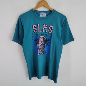 Puede incluir: Camiseta de color turquesa con un gráfico que presenta la palabra "SLRS" en letras rosas con burbujas sobre un diseño tiki estilizado. El tiki es rosa y negro, sobre un fondo azul con llamas. El texto "GDC SAILORS" está debajo.