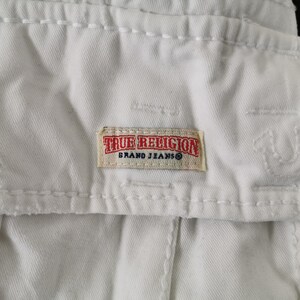 May include: Close-up of white denim fabric with visible stitching and a rectangular True Religion Brand Jeans label. The label is beige with red and black text.