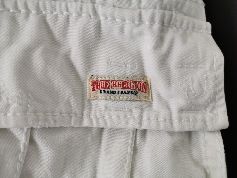 May include: Close-up of white denim fabric with visible stitching and a rectangular True Religion Brand Jeans label. The label is beige with red and black text.