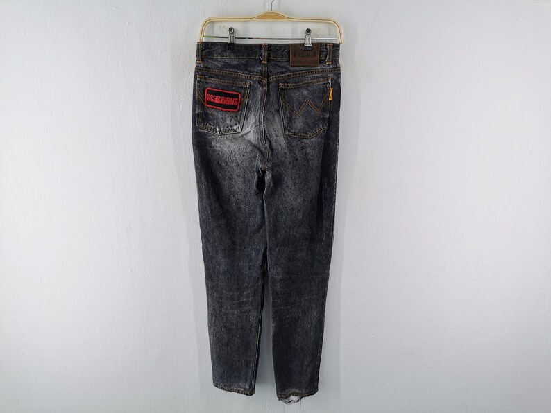 May include: Black acid-wash denim jeans featuring a rectangular red patch with the text SCORPIONS on the left back pocket, a brown leather brand label on the waistband, and a signature W-shaped stitching detail on the right back pocket.