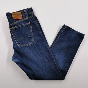 May include: Dark blue denim jeans with a classic five-pocket design. Features a brown leather patch with the brand name "EDWIN" on the back waistband. The back pocket has a unique zigzag stitch detail. The jeans are laid flat.